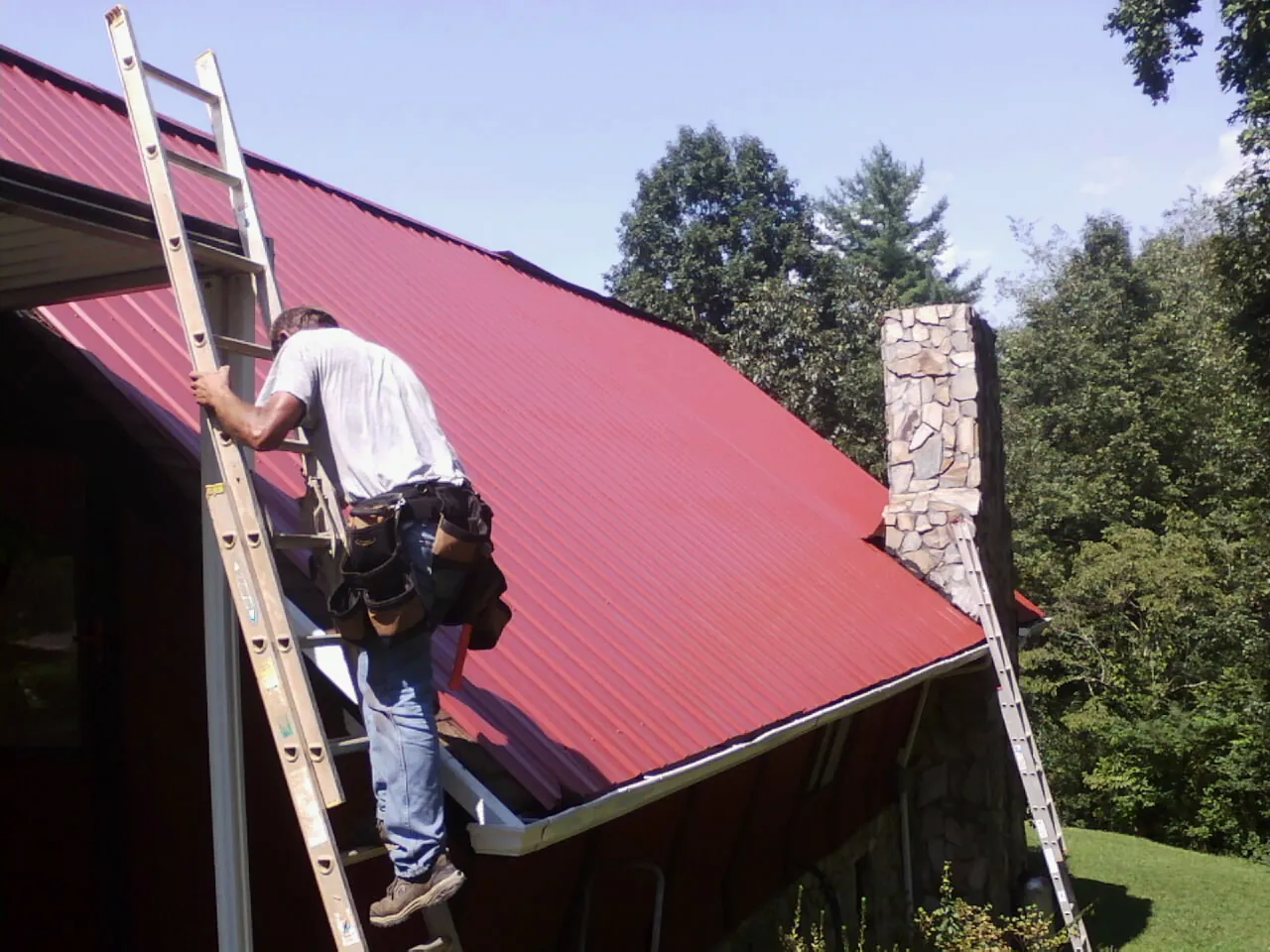 Roofer on a ladder beside a red metal roof and stone chimney — Soffit & Fascia Repair by Roofing Brothers in Jersey Mills